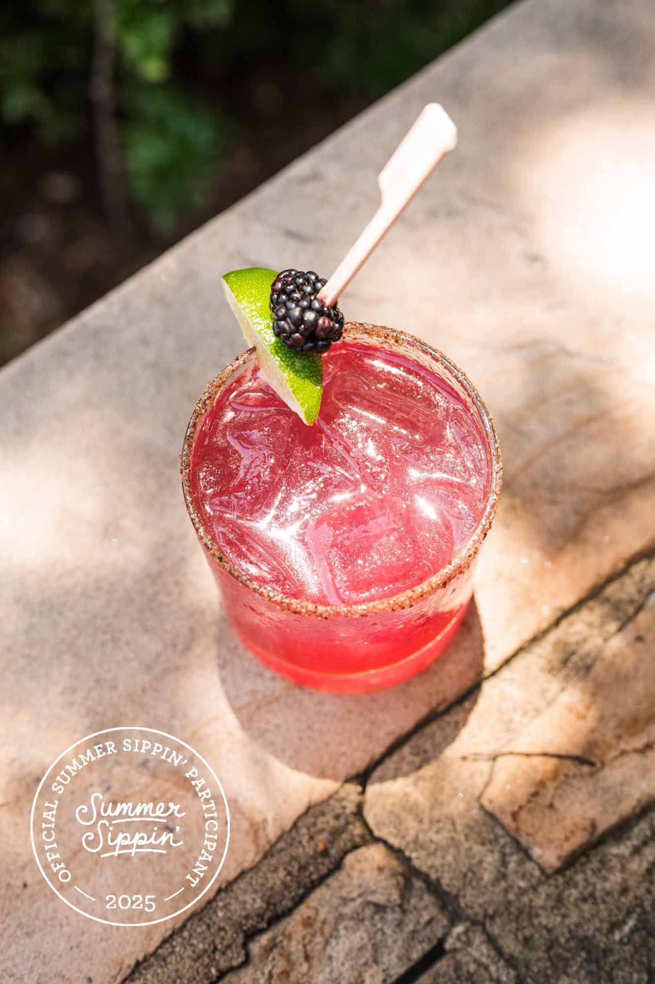 Cocktail with red liquid, lime, and blackberry garnish, salt rim, outdoors on a stone surface.
