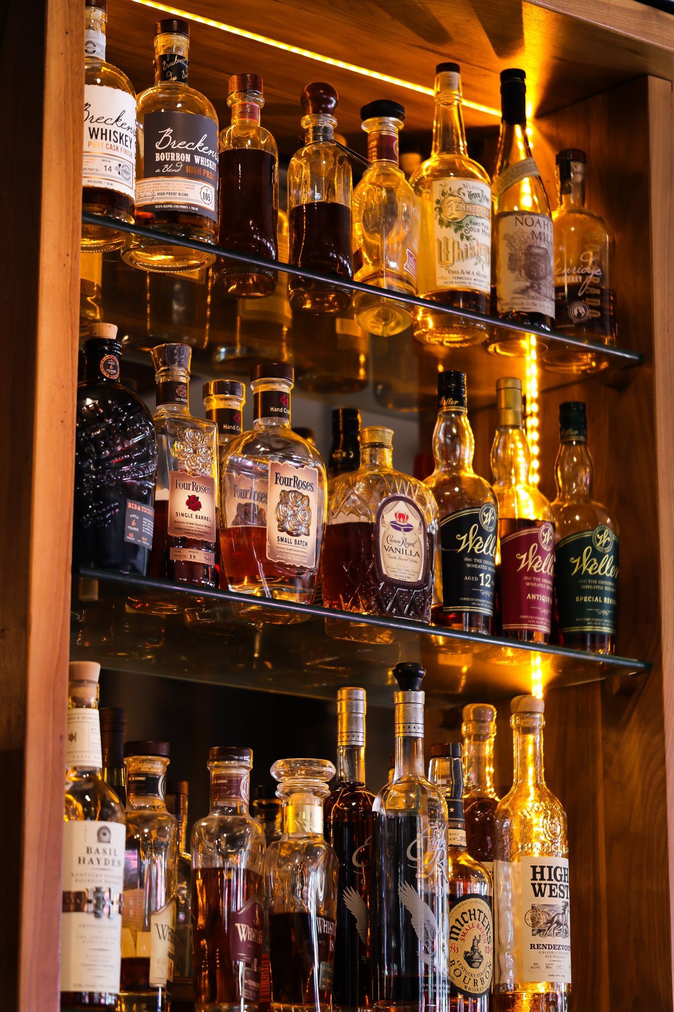 Liquor bottles on lit wooden shelves. Warm lighting highlights varied bottle shapes and colors.