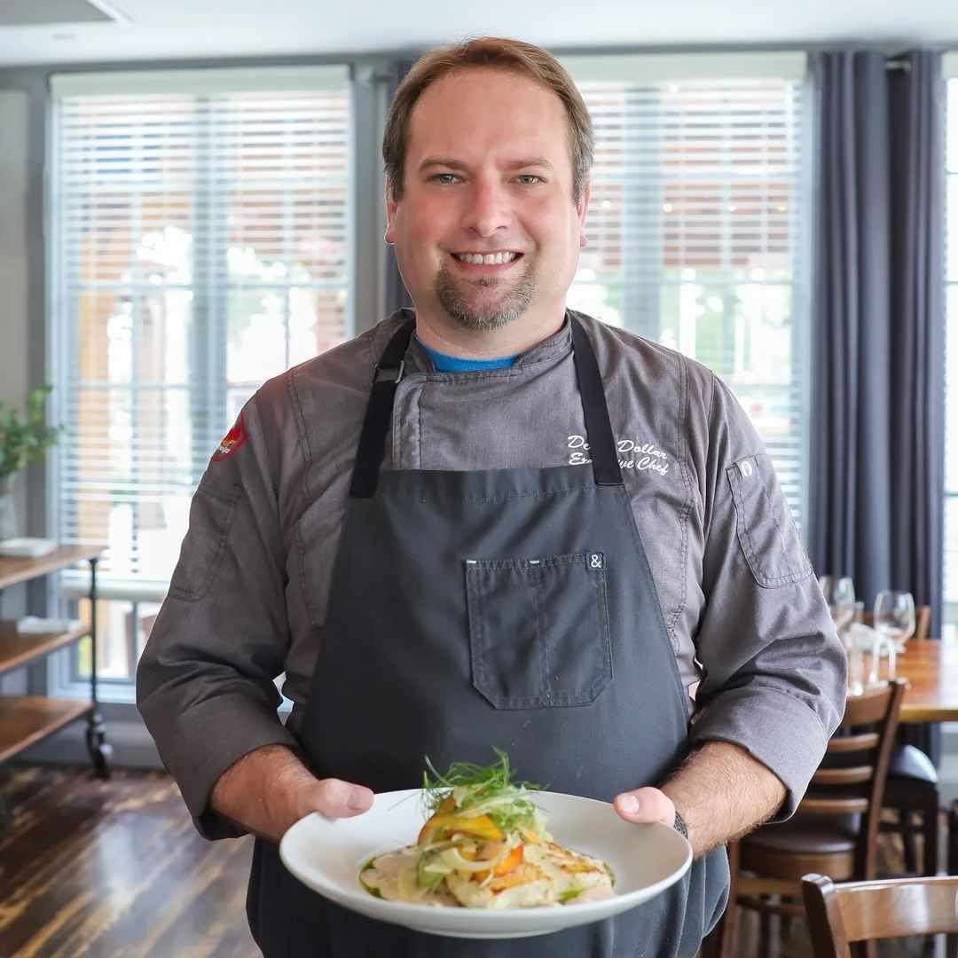 Chef holding a plate of food, smiling. Gray apron, dining room setting.