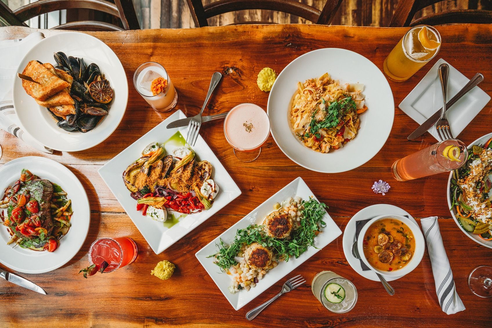 Overhead shot of a wooden table laden with various dishes and cocktails at a restaurant.