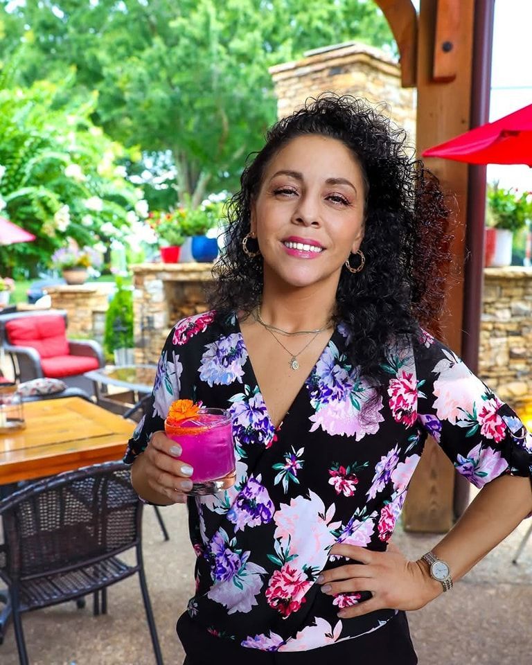 Woman outdoors holding a pink cocktail, smiling, wearing floral top, restaurant patio in background.