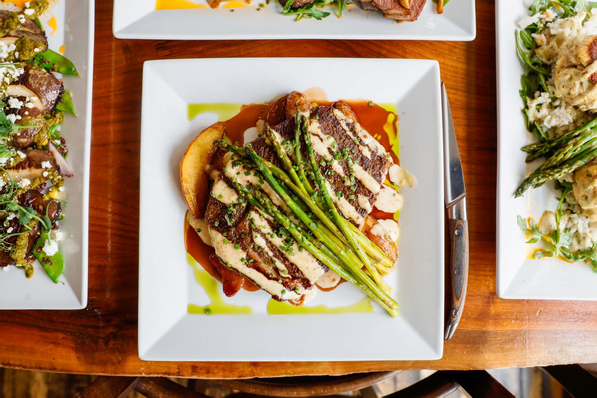 Plate of food with steak, asparagus, and sauce, on a wooden table. Other plates visible.