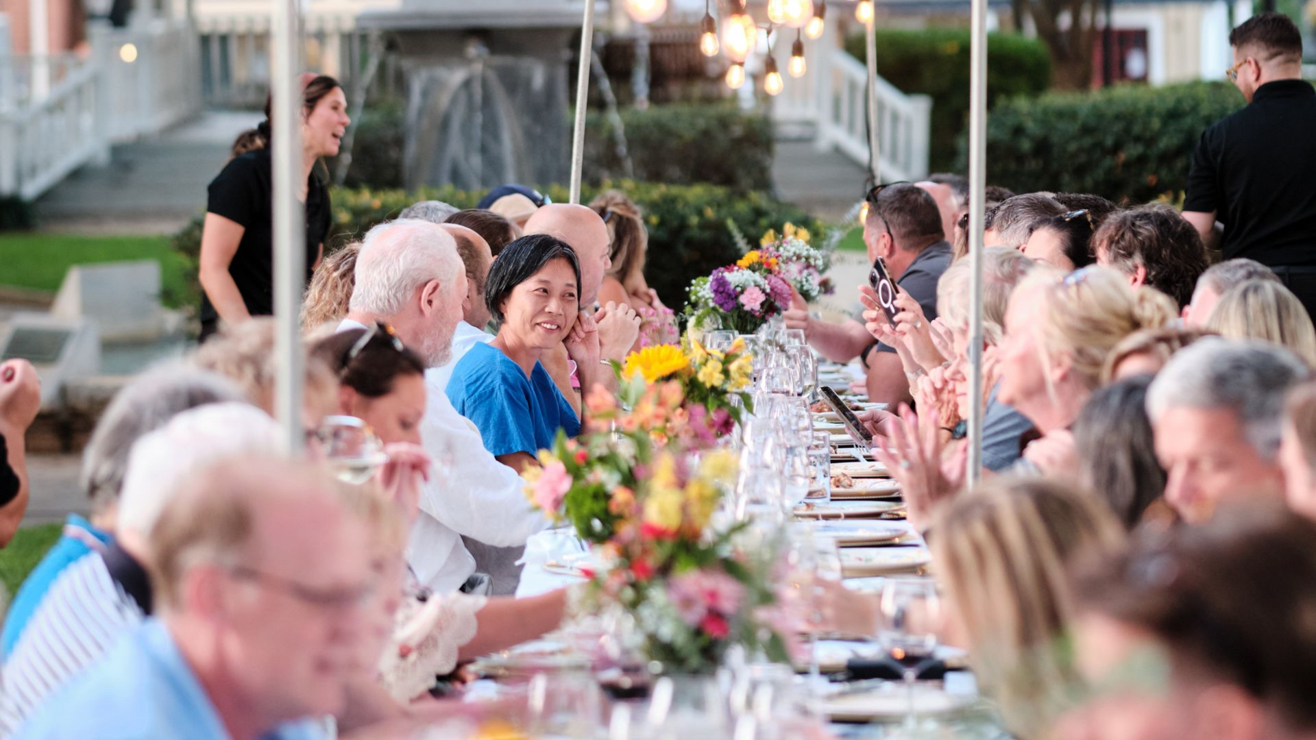 People seated at a long table for an outdoor dinner party, colorful flowers, overhead lights, server.