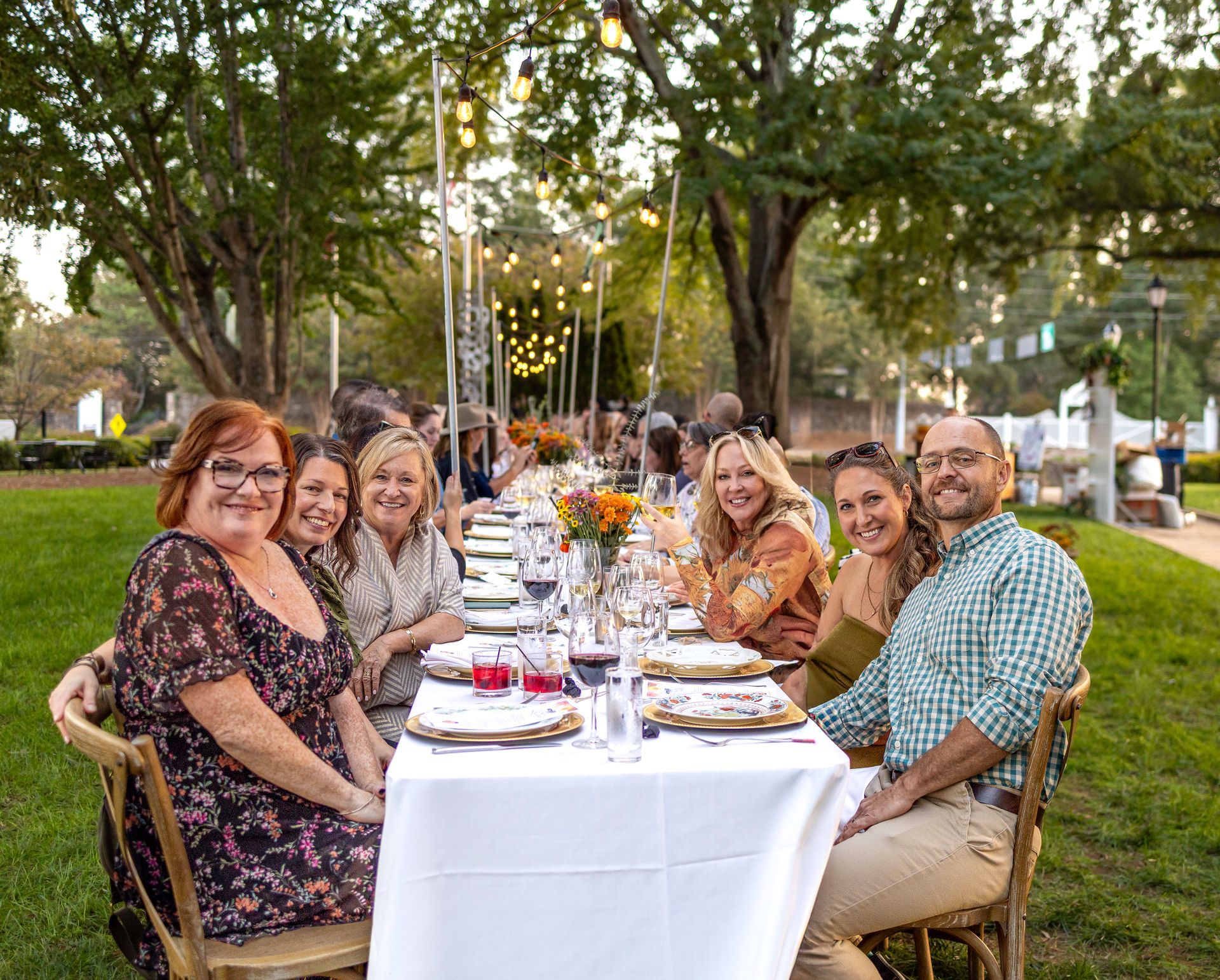People seated at a long table set for a meal outdoors. Trees and string lights are overhead.