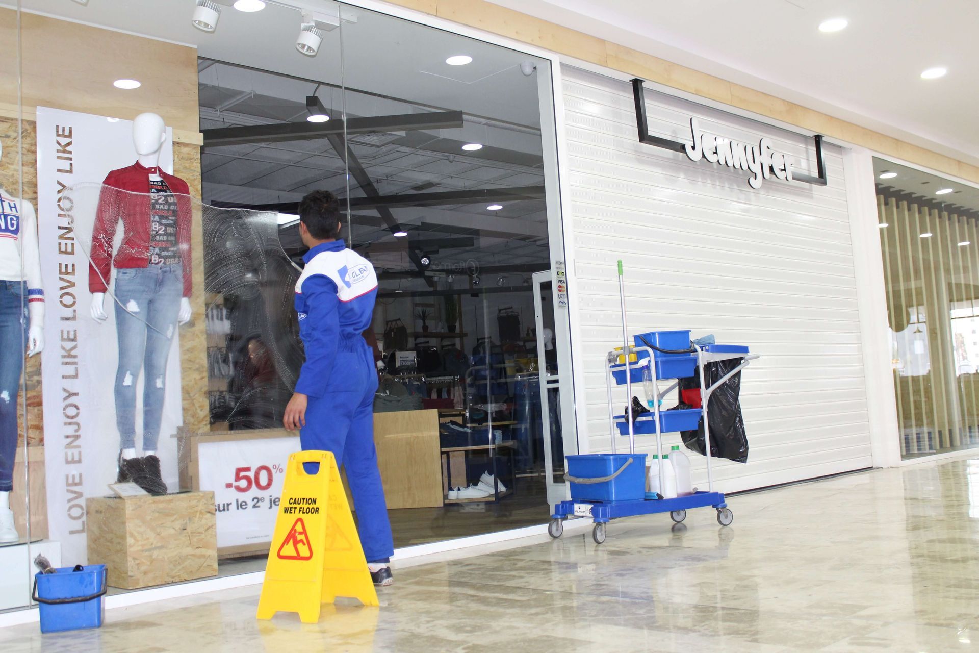 A person in blue overalls stands near a wet floor sign outside a clothing store.