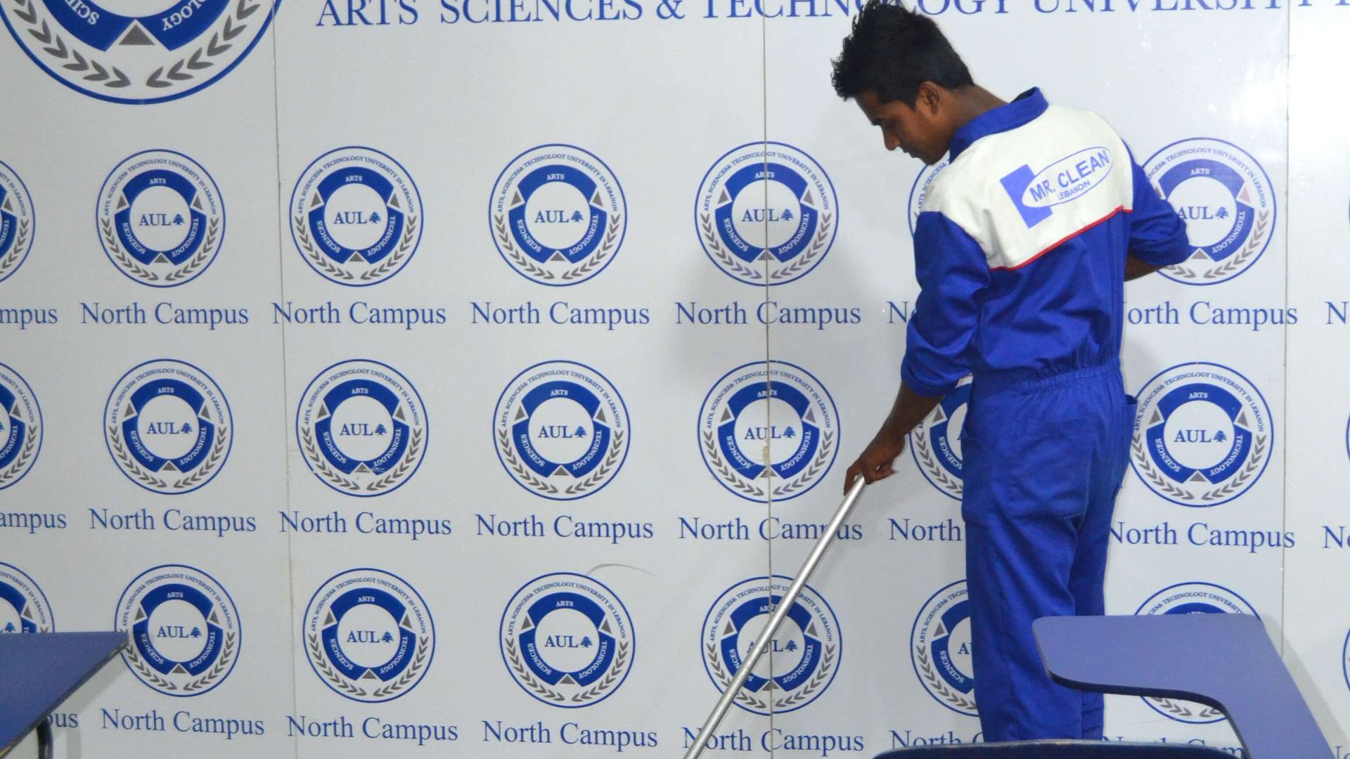 Man in blue uniform cleaning floor, wall with repeated university logo in background.