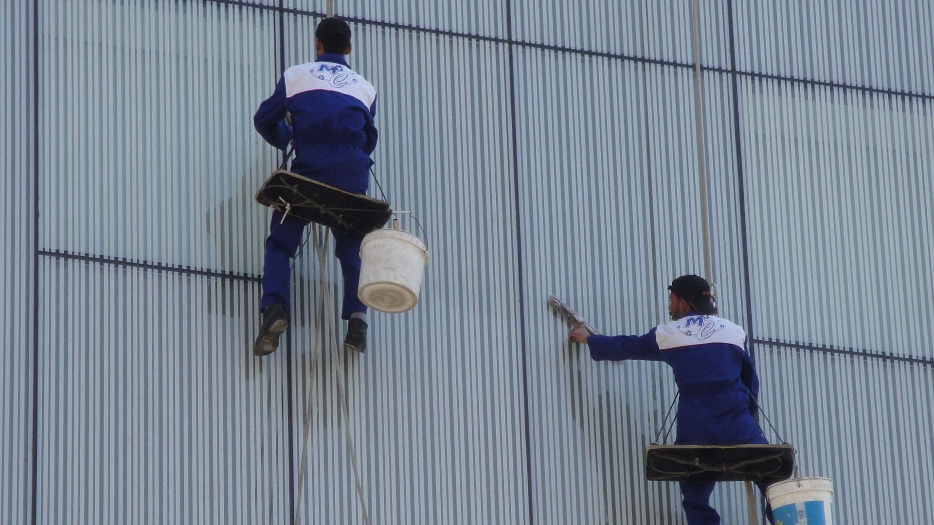 Two workers in blue suits on scaffolding, washing the outside of a tall building.
