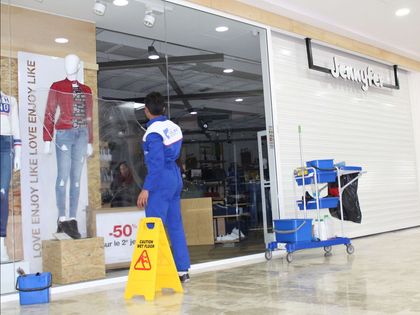 Man cleaning a storefront after glass damage, with a wet floor sign and cleaning cart in a mall.