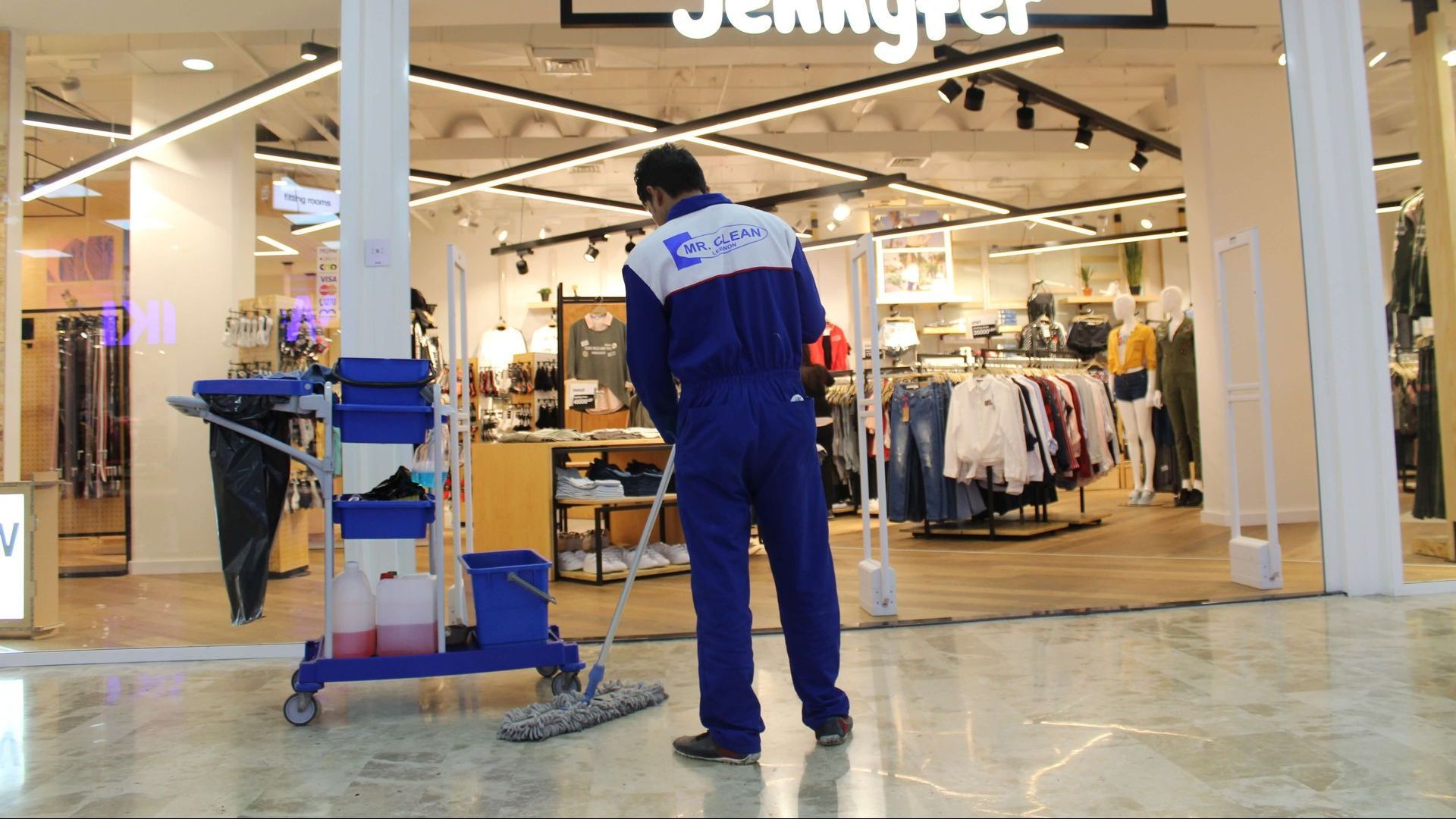 A person in blue uniform mops a store floor in a mall; a cleaning cart is nearby.