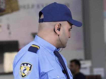 Security guard in blue uniform and cap, wearing an earpiece, looking to the side.