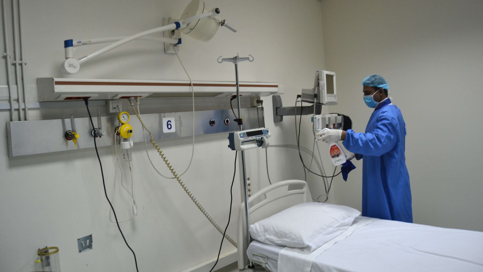 A medical professional in blue scrubs adjusts equipment in a hospital room with an empty bed.