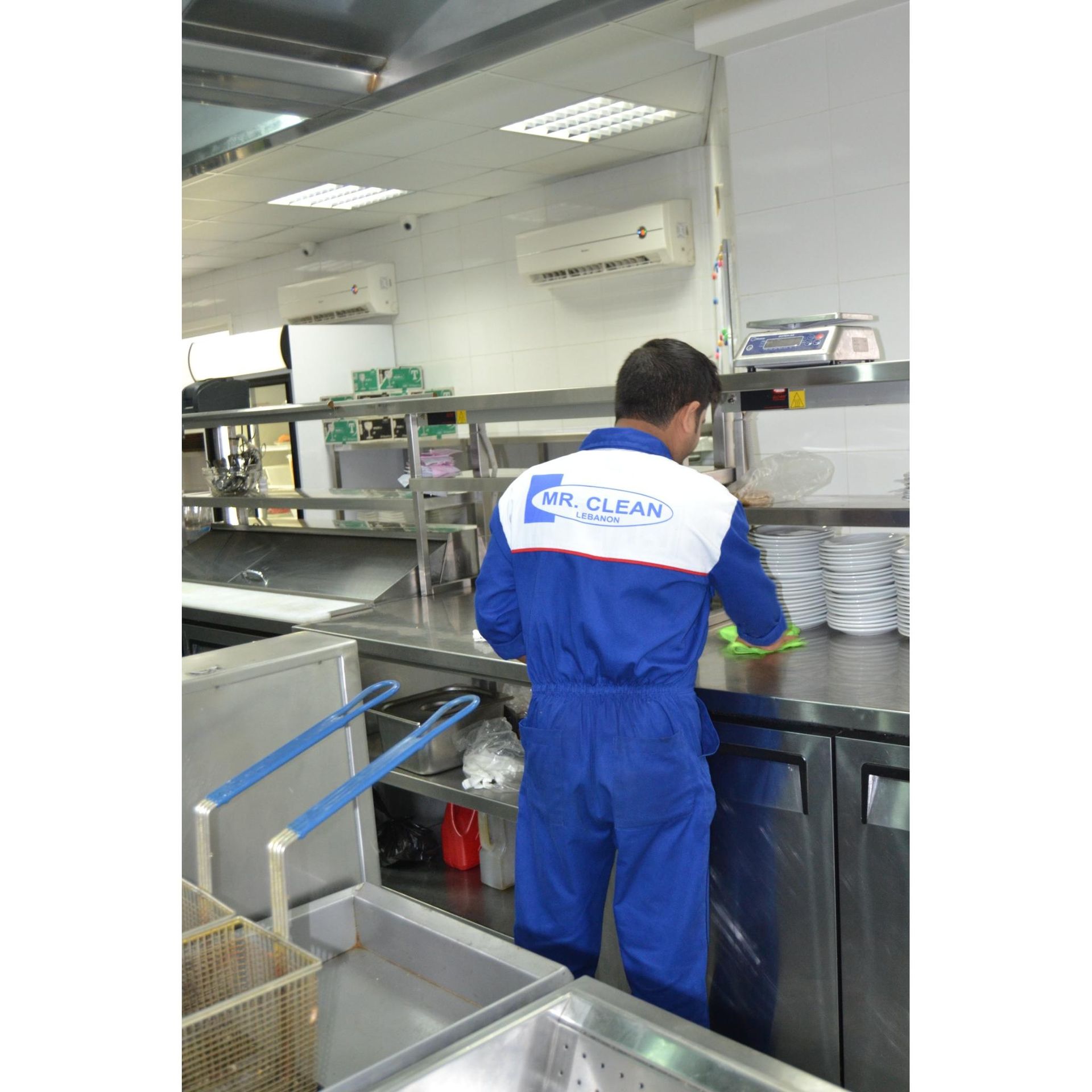 Person in blue uniform cleaning a commercial kitchen. Stainless steel counters and appliances.