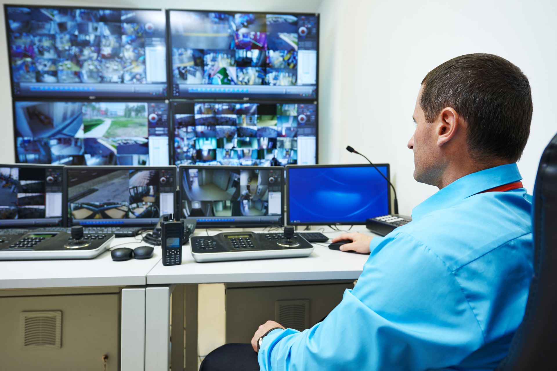 Man in blue shirt monitors security cameras at a desk with multiple screens.