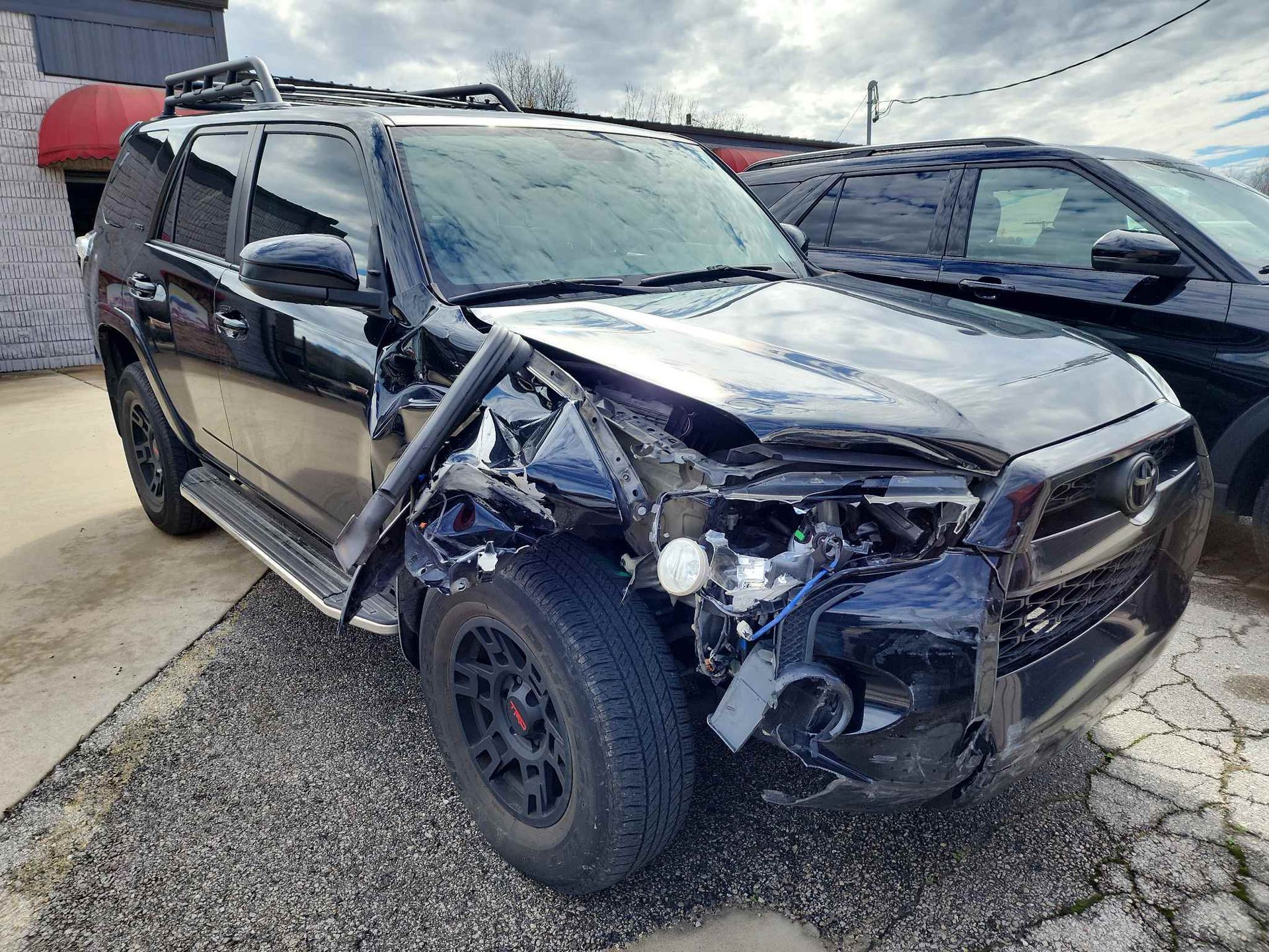 Black Toyota SUV with significant front-end damage parked outside a building on a cloudy day.