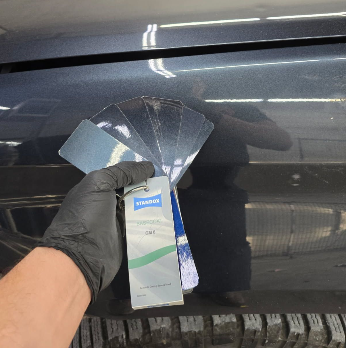 Gloved hand holding a paint sample fan beside a dark car door, likely for color matching.