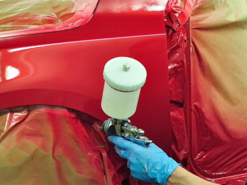 Gloved hand sprays red car body panel with paint gun and white paint cup
