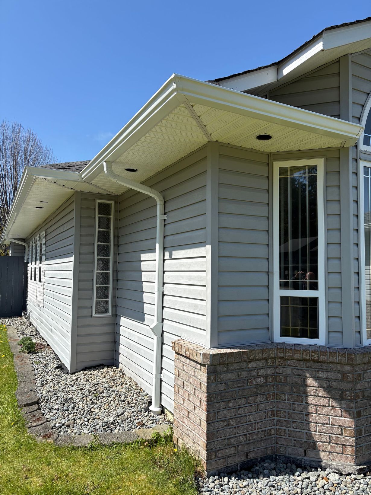 Gray house exterior with white trim, gutters, and downspout; brick base, rocky landscaping, blue sky.