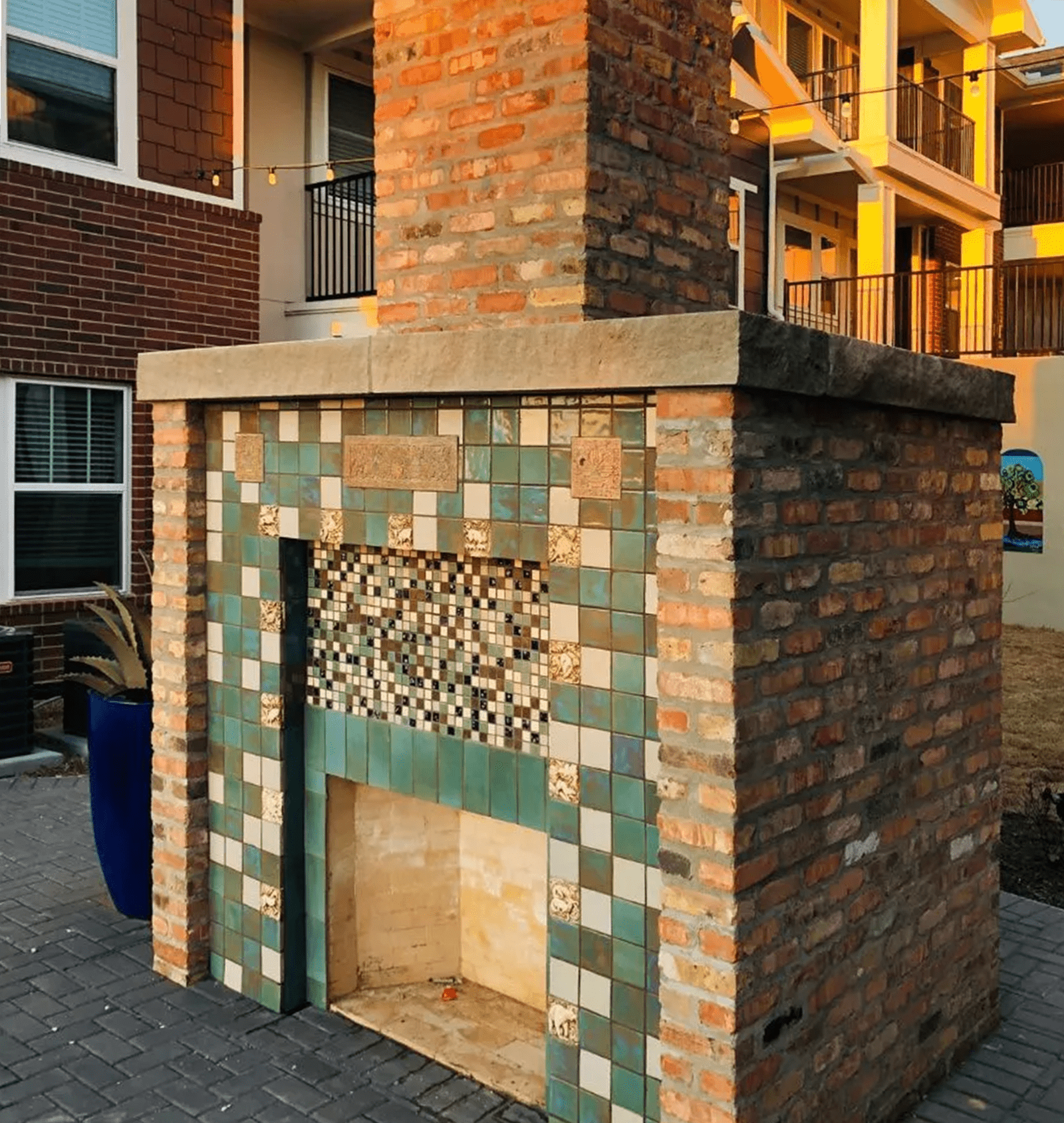 Brick outdoor fireplace with green and white tiles, in front of apartments.