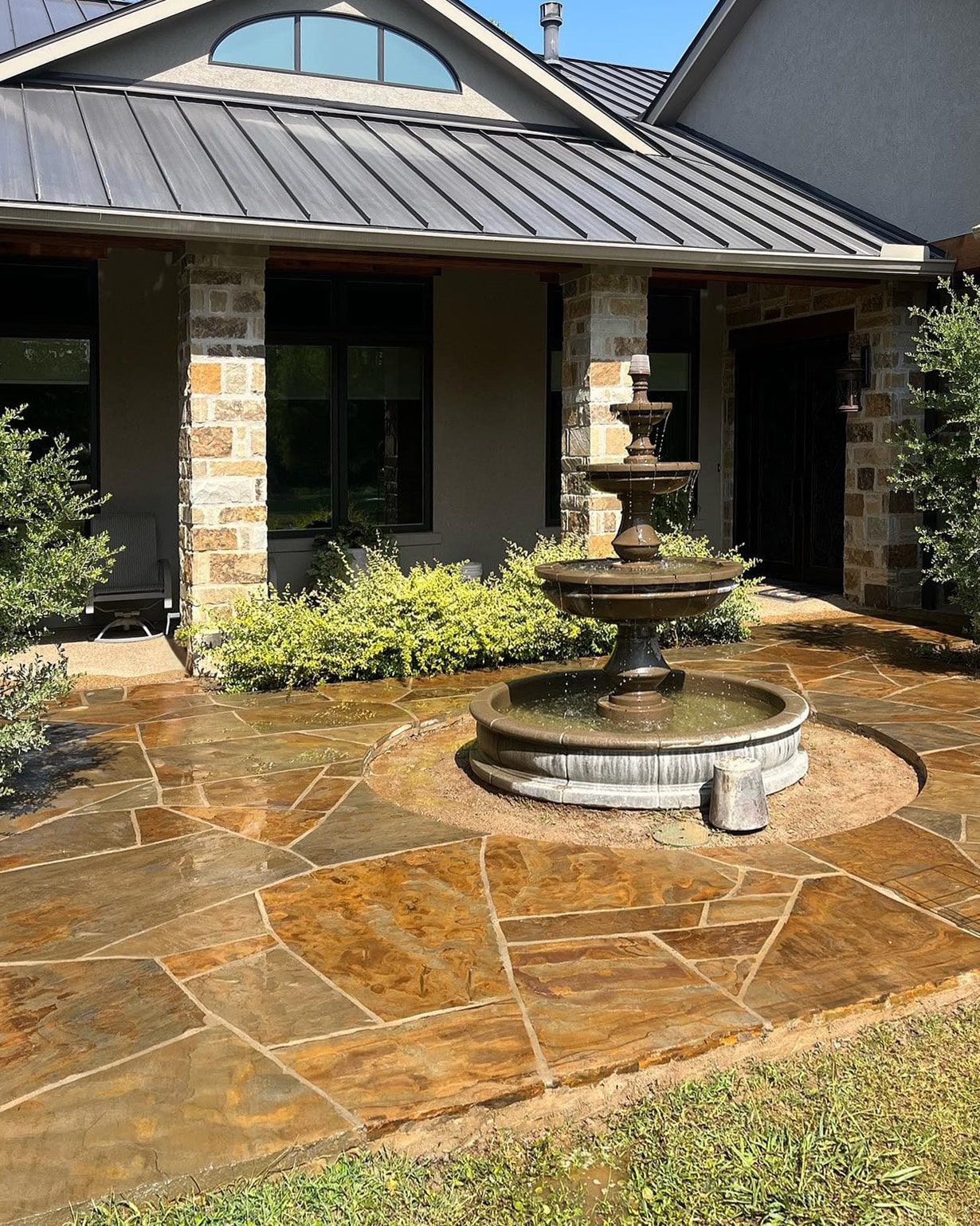 Stone patio with a fountain in front of a building with stone pillars and metal roof.