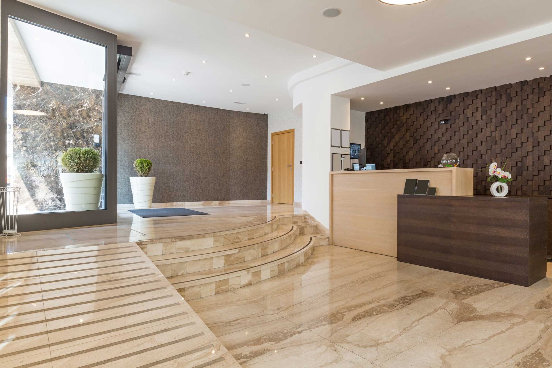 Modern hotel lobby with marble floors, curved steps, front desk, and potted plants.