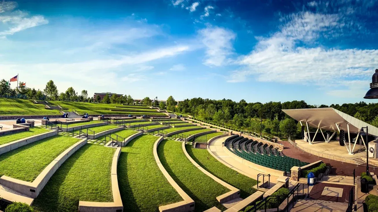 Outdoor amphitheater with tiered, grassy seating, stage, and blue sky.