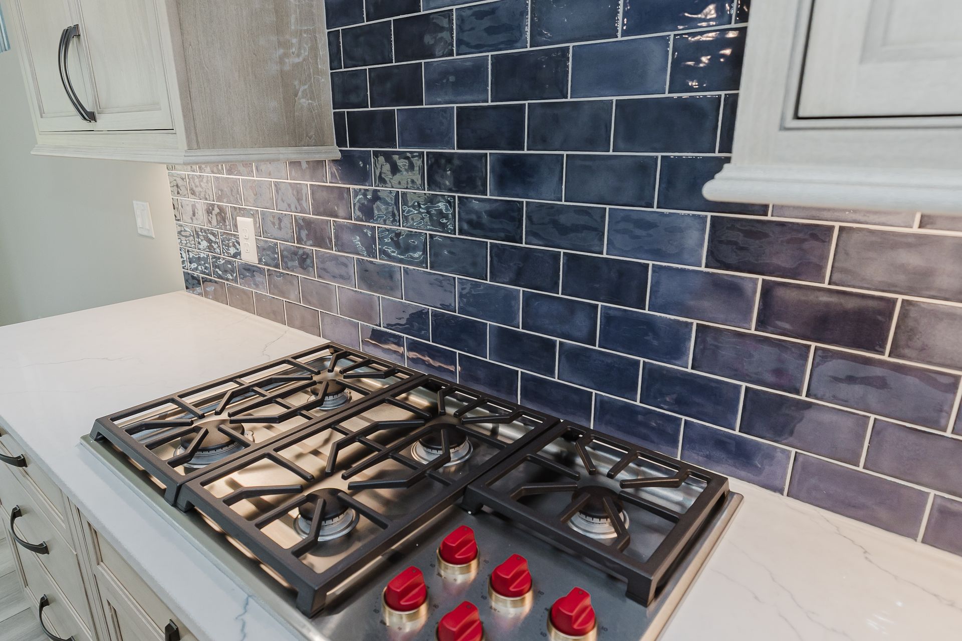 Gas stove with red knobs on a white countertop in the Humphrey Bogart model, blue tiled backsplash, and white cabinets.