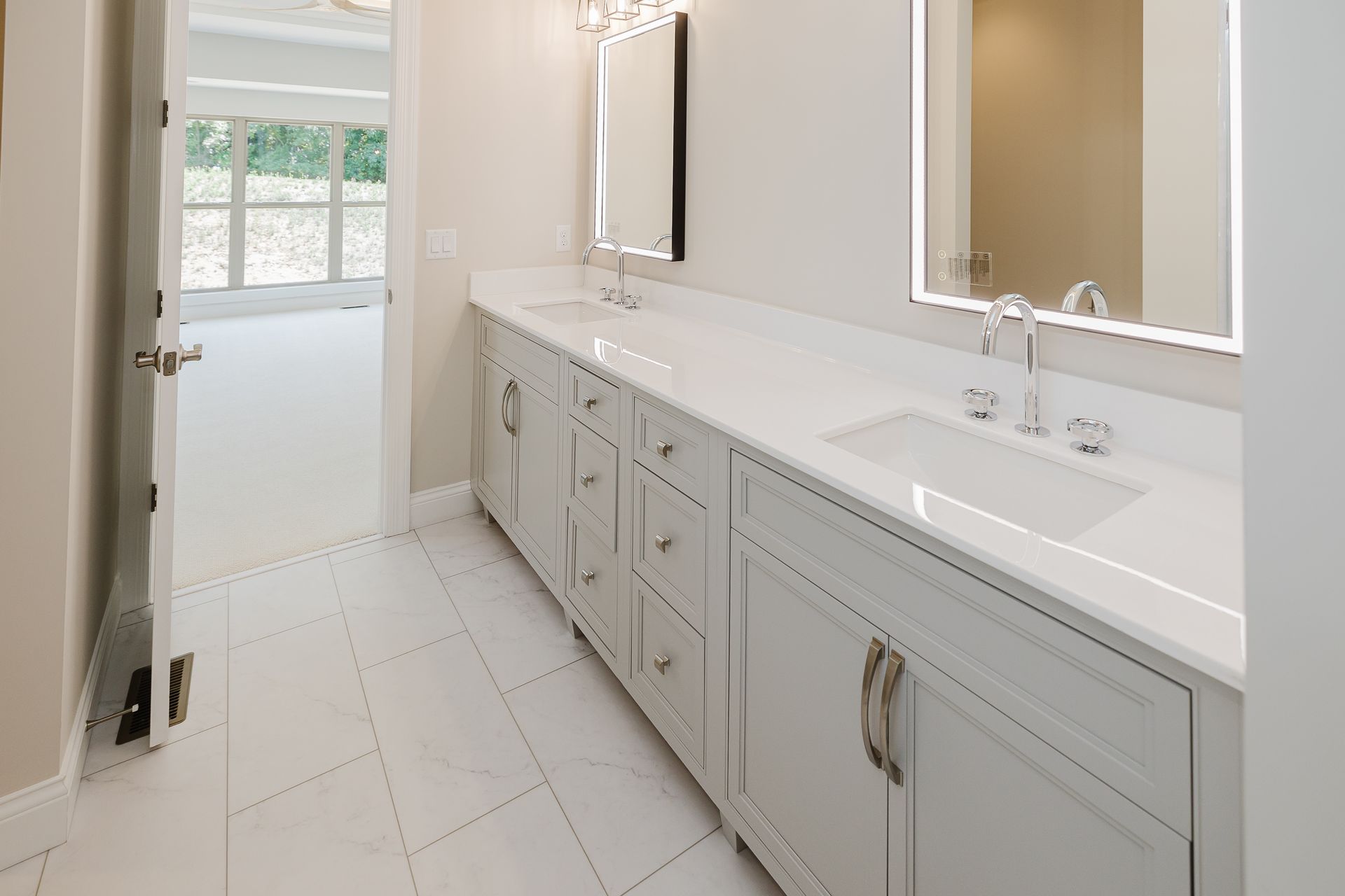 White bathroom of the Humphrey Bogart model with double vanity, sinks, mirrors, and door leading to a room with window.