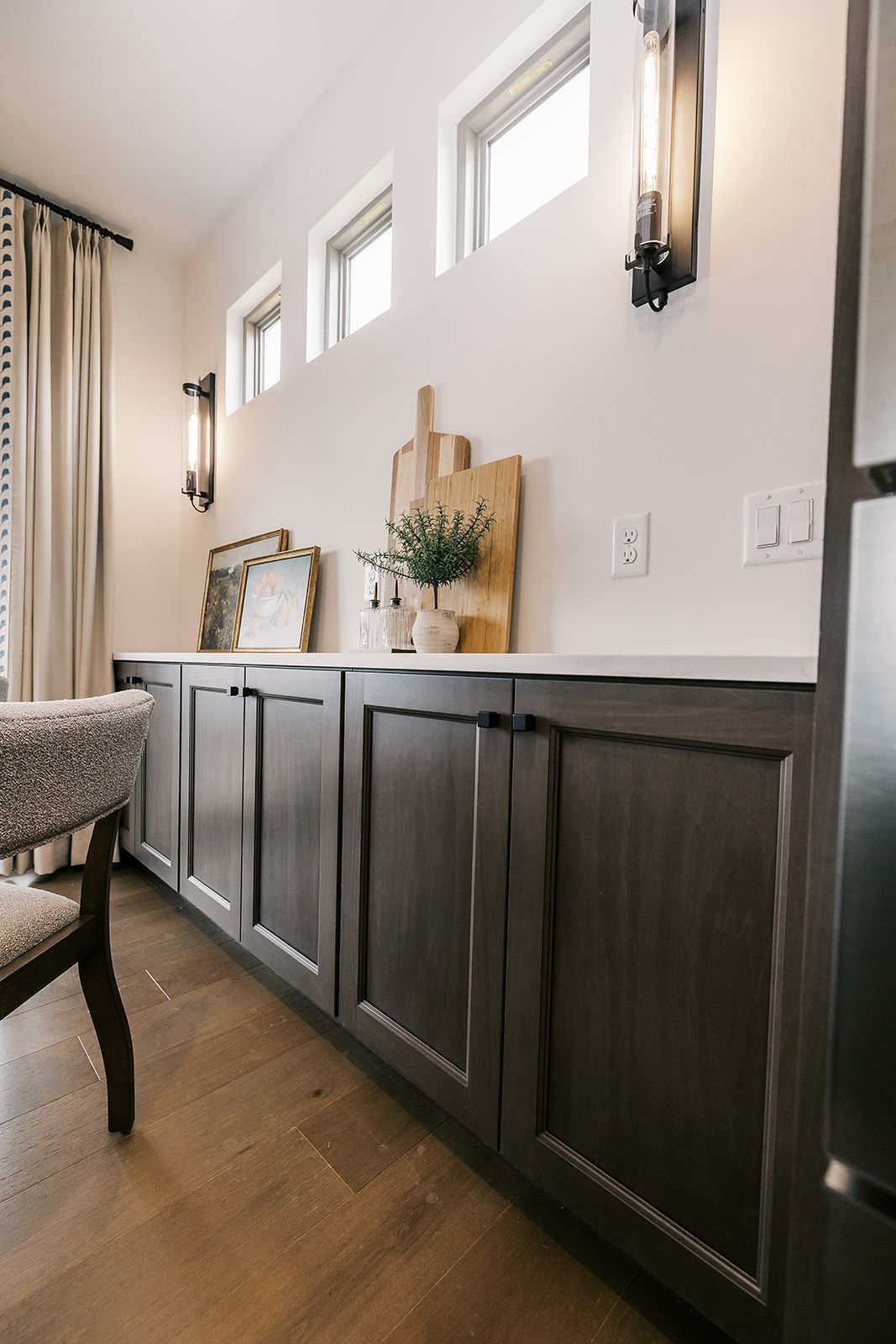 Dark wood cabinets in the Cary Grant model with white countertop, windows above. Sconces and decor adorn the wall.