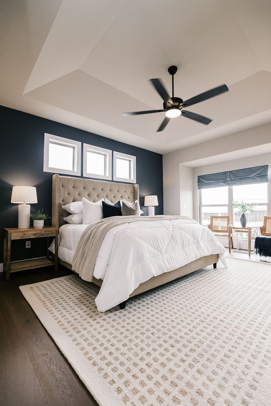 Bedroom in the Cary Grant model with navy accent wall, tufted headboard, and cream rug. Ceiling fan overhead.