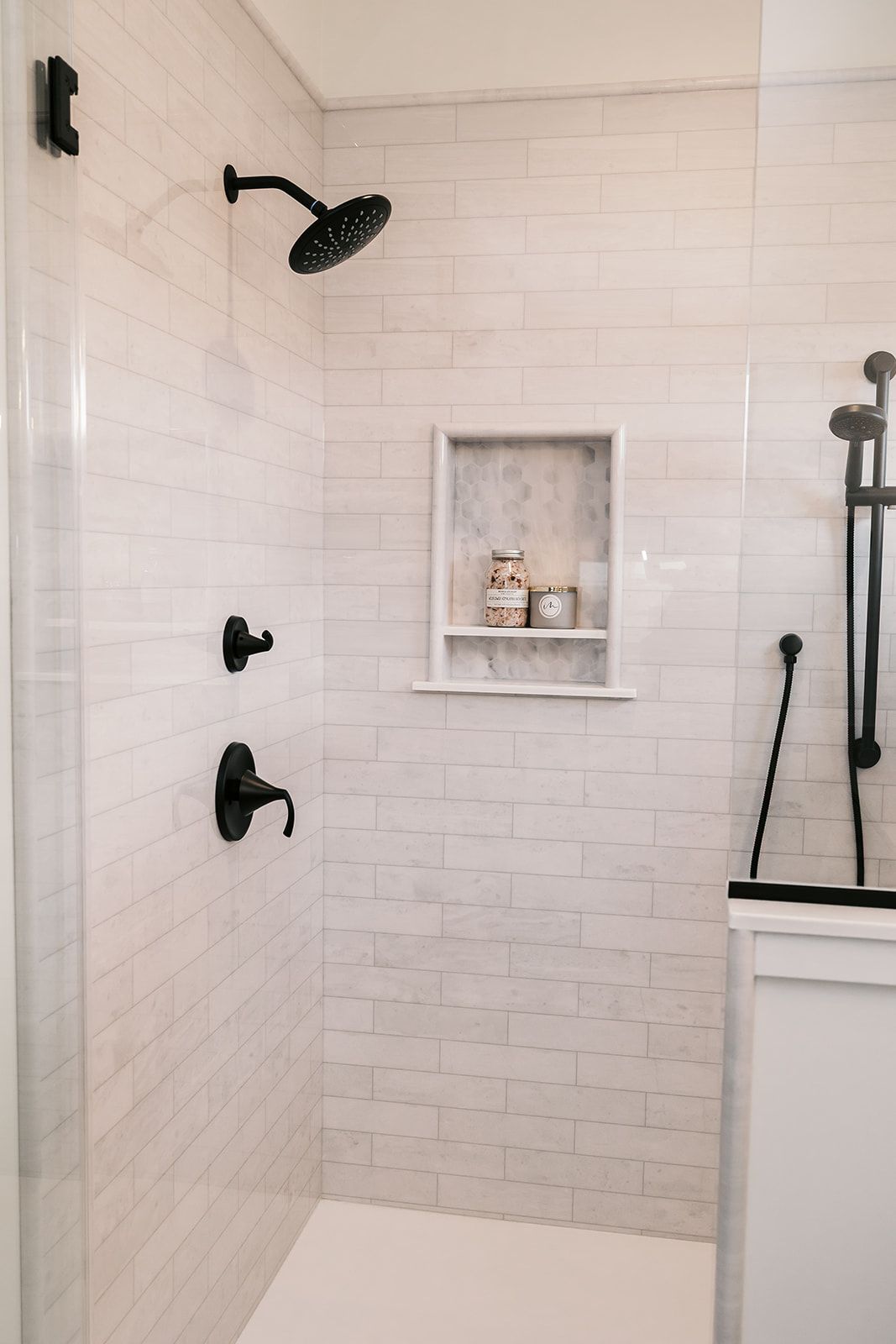 White-tiled shower in the Cary Grant model with black fixtures and a recessed shelf. A glass door is on the left.