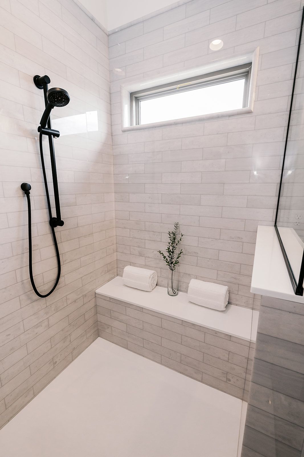 Modern white-tiled shower  in the Cary Grant model with black fixtures, bench, small window, and towels.