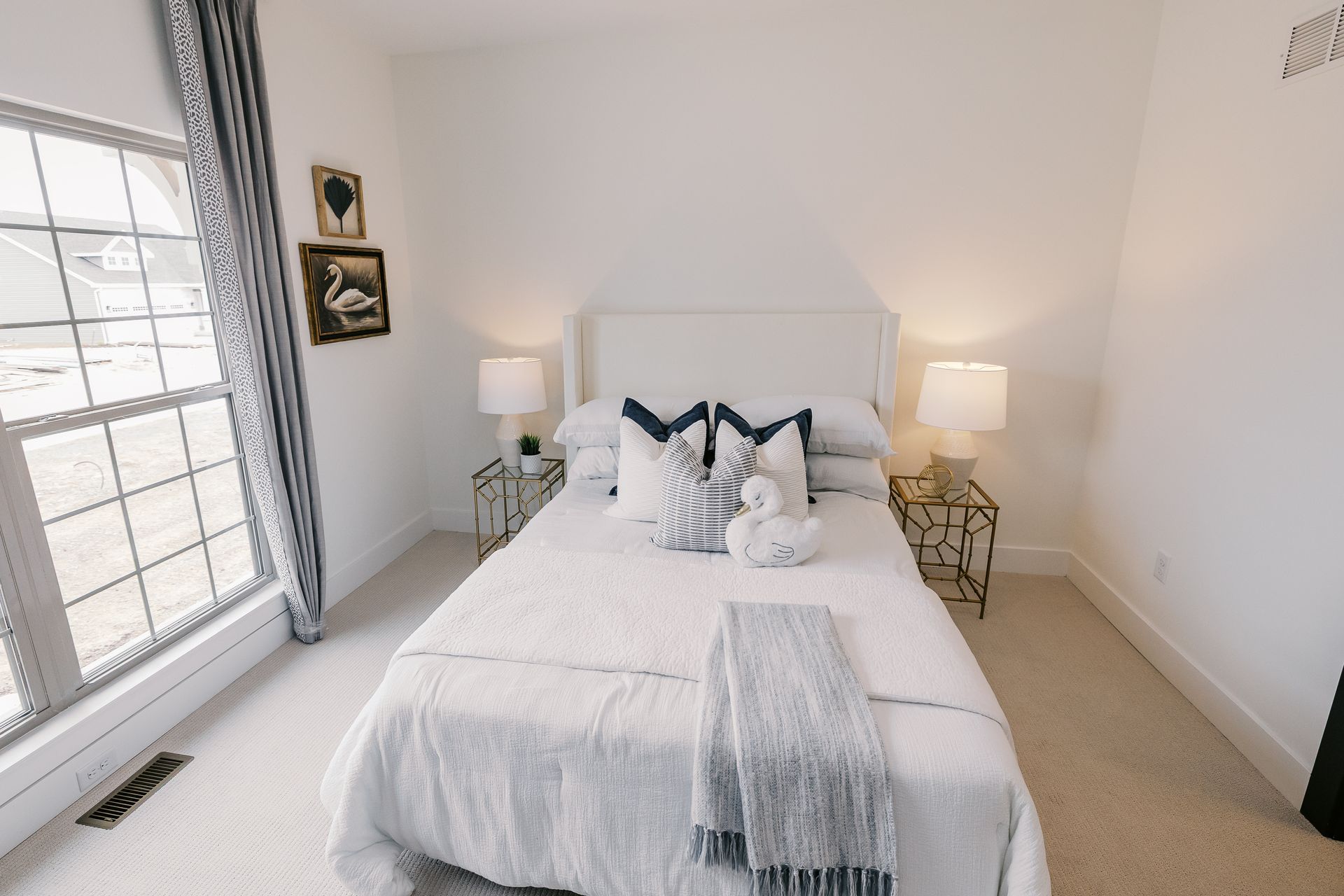 Bedroom in the Cary Grant model with a white bed, decorative pillows, lamps, and a window with gray curtains.