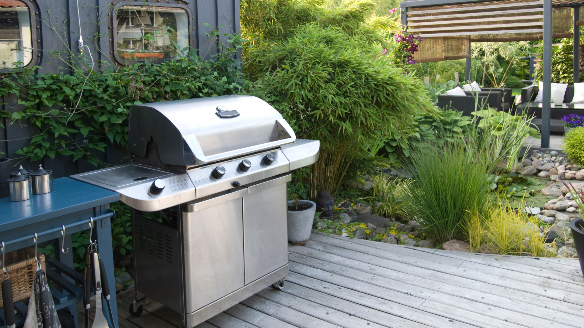 Stainless steel gas grill on a wooden patio surrounded by greenery and garden plants