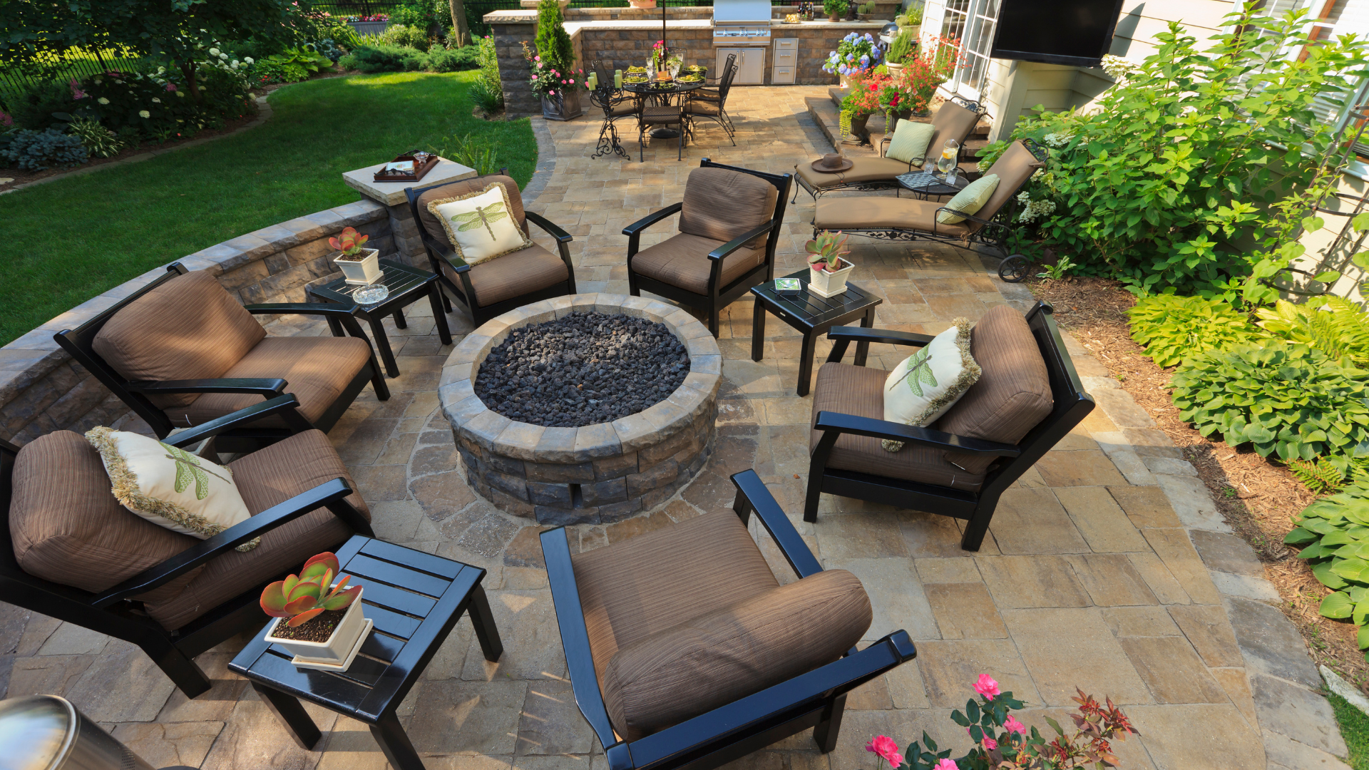 Outdoor stone patio with a circular fire pit and cushioned brown chairs arranged around it