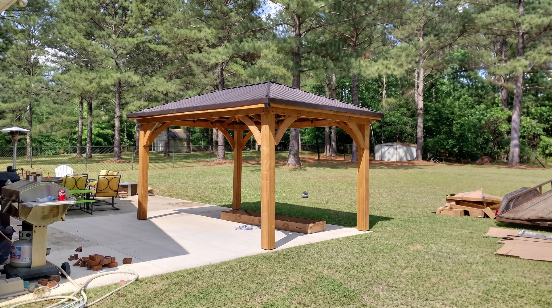 Wooden gazebo with dark roof on a concrete patio in a grassy backyard, surrounded by trees.