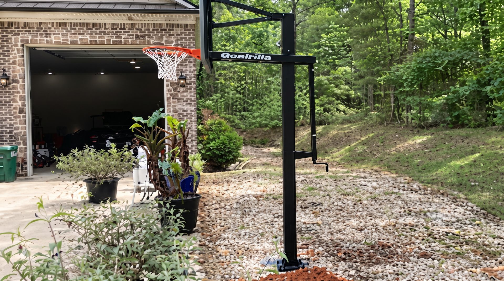Portable basketball hoop beside an open garage, with a gravel driveway and trees in the background