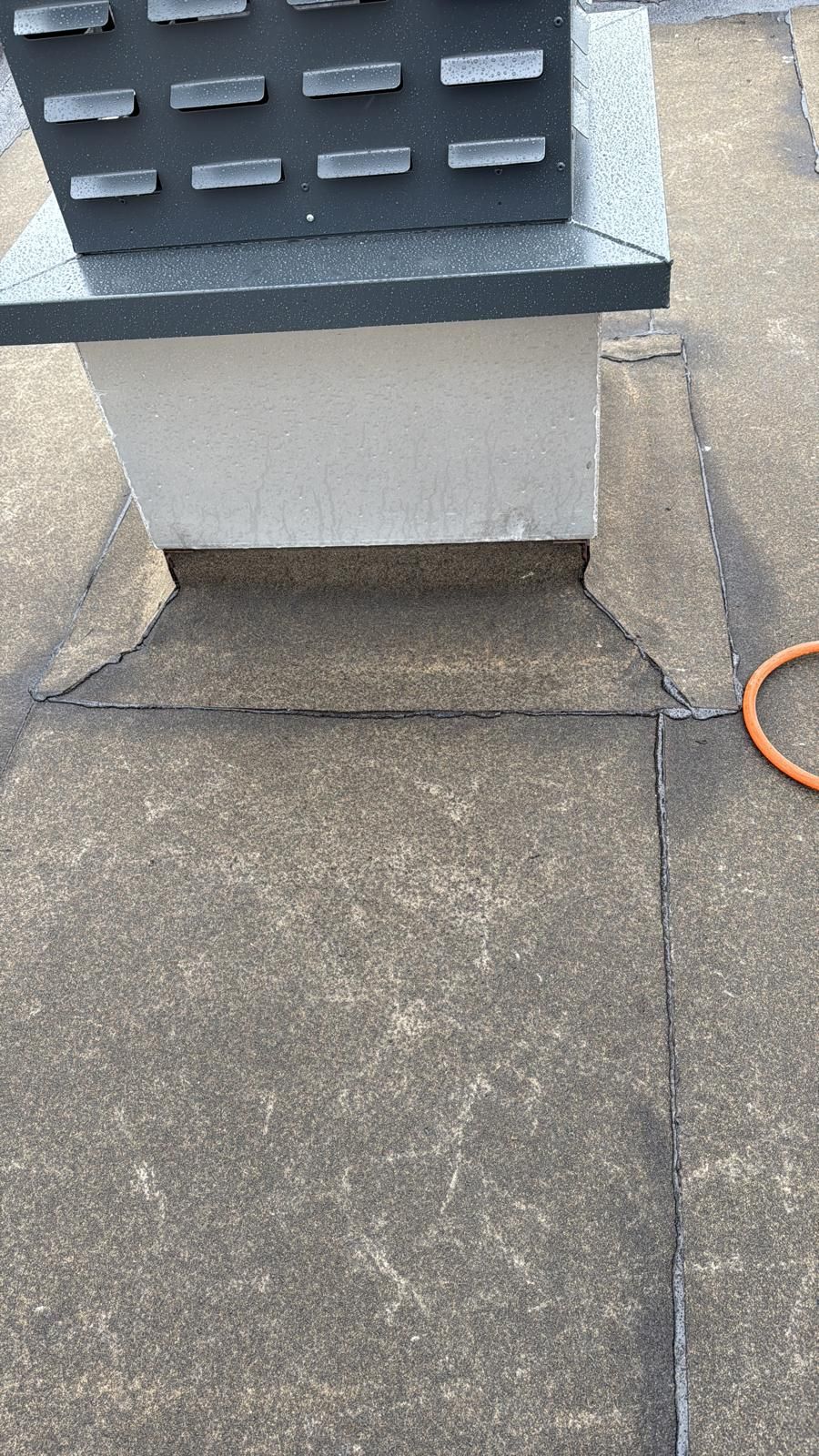 Een grijze schoorsteenvoet op een plat, betonkleurig dak met een beschermende metalen kap en zichtbare naadlijnen.