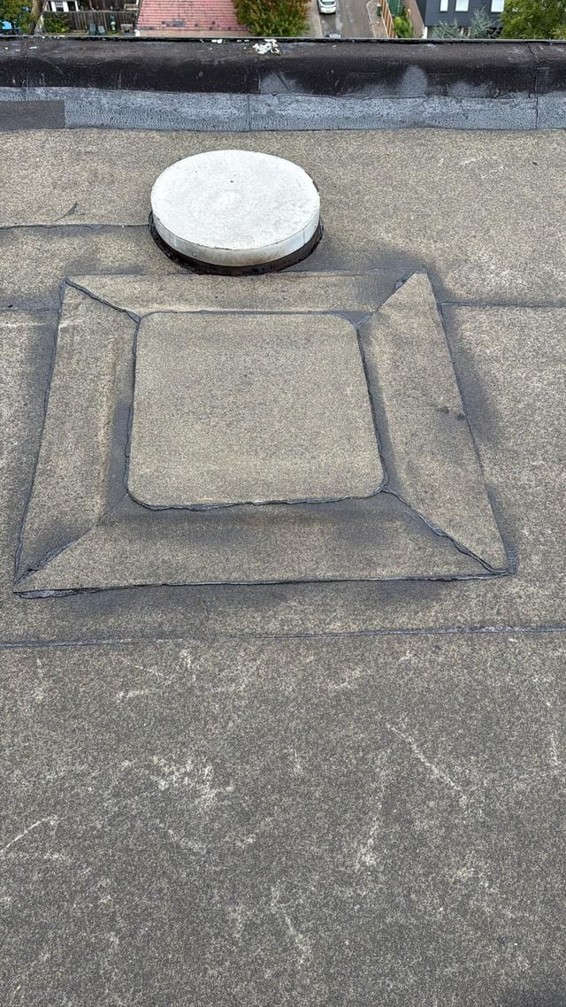 Een grijs, vlak dakoppervlak met een vierkant, verhoogd gedeelte en een ronde, lichtgekleurde ventilatiekap.
