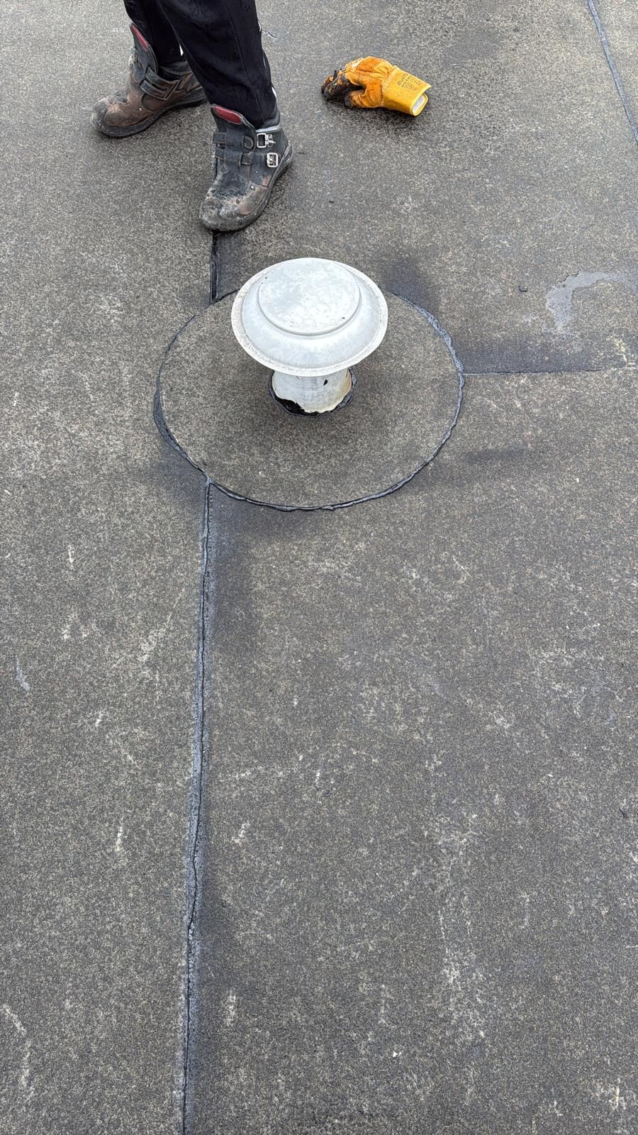 Een persoon staat op een plat, grijs dak naast een ronde, witte ventilatiekap in het midden van een stuk dakbedekking.