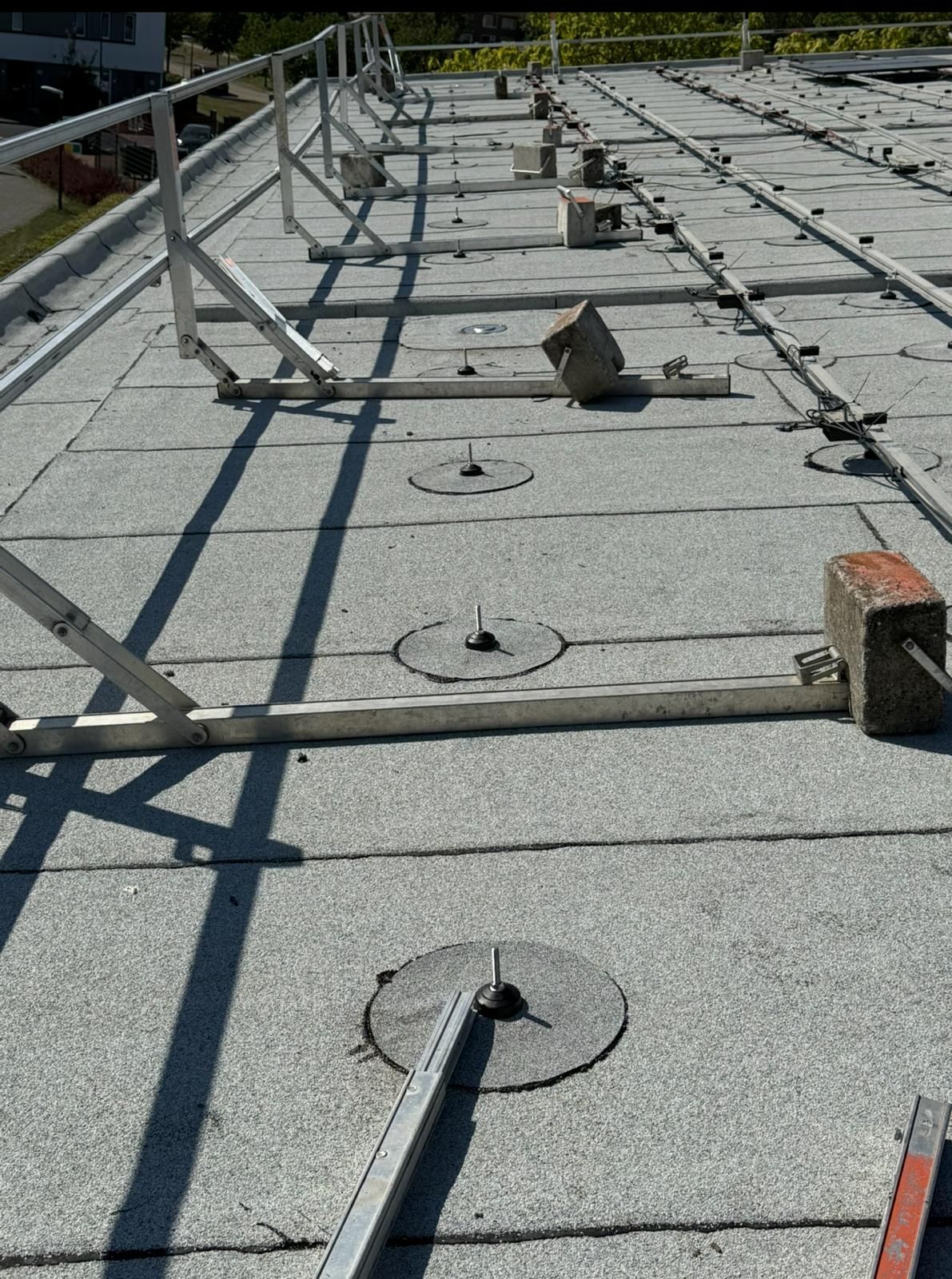 Een plat grijs dak in aanbouw met rijen metalen framebalken en ankerplaten die verankerd zijn met betonblokken.