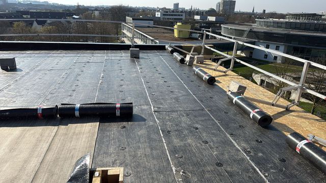 Zwart, uitgerold dakmembraan op een plat dak van een bouwplaats, met metalen hekwerk op de achtergrond.
