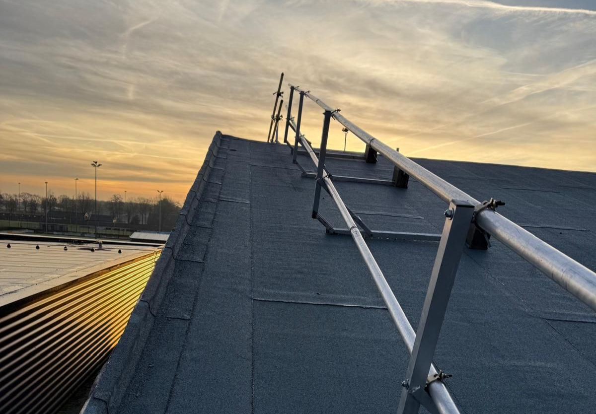 Een metalen veiligheidsleuning loopt langs het schuine, donkergrijze dakoppervlak bij zonsondergang.