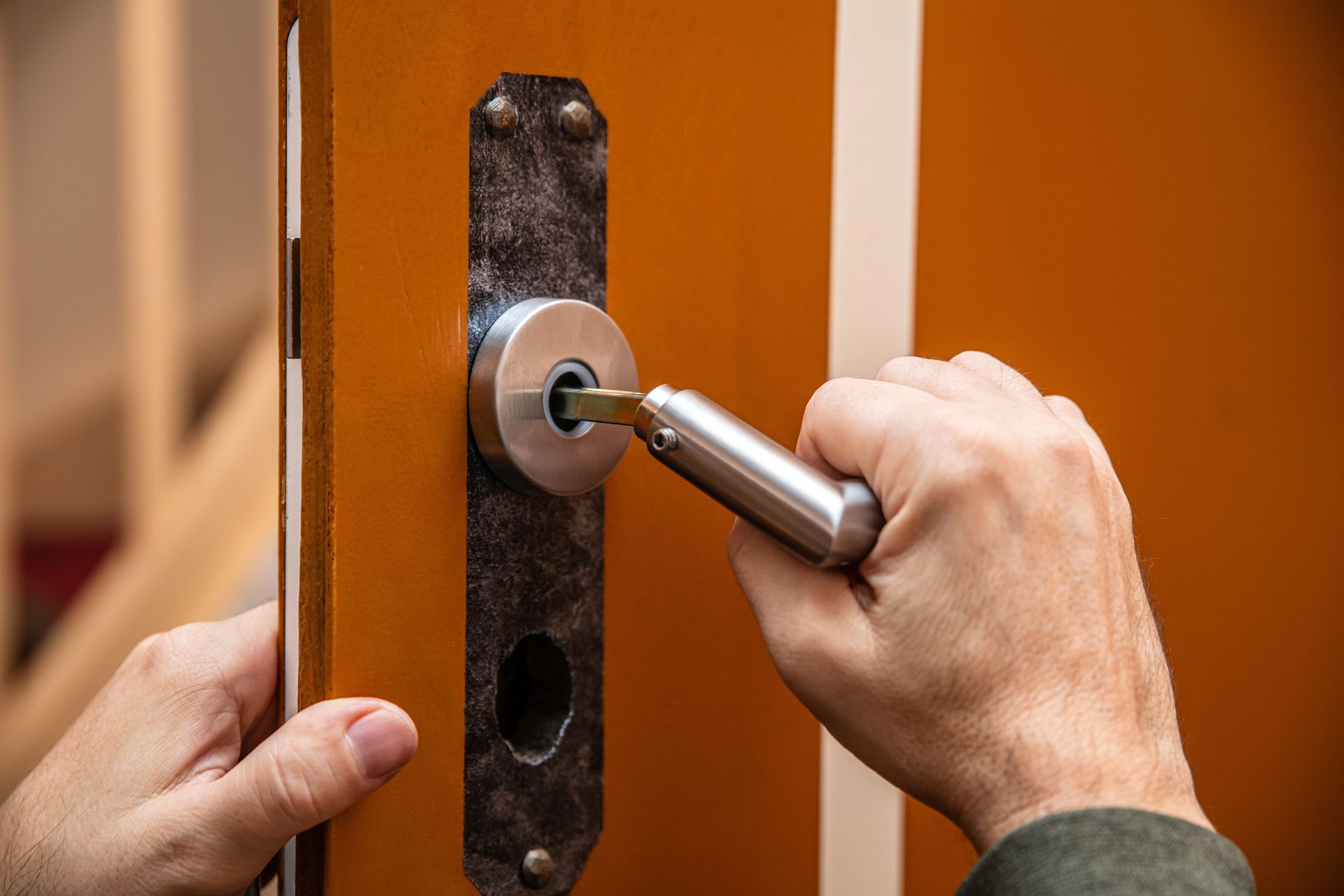 Hands using a screwdriver to remove a door lock cylinder from a black metal lock plate on an orange door