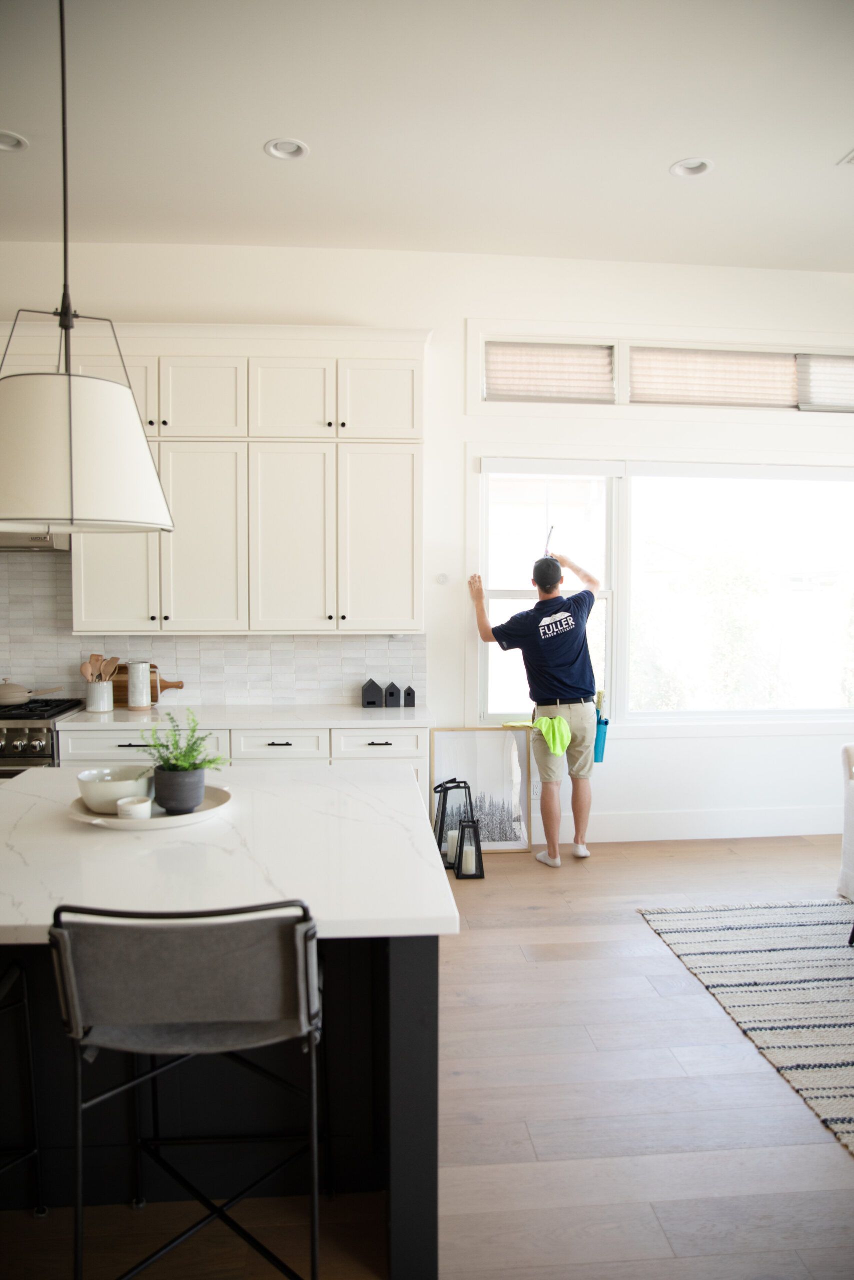 Bright kitchen with white cabinets; person standing by the window reaching up to adjust blinds.