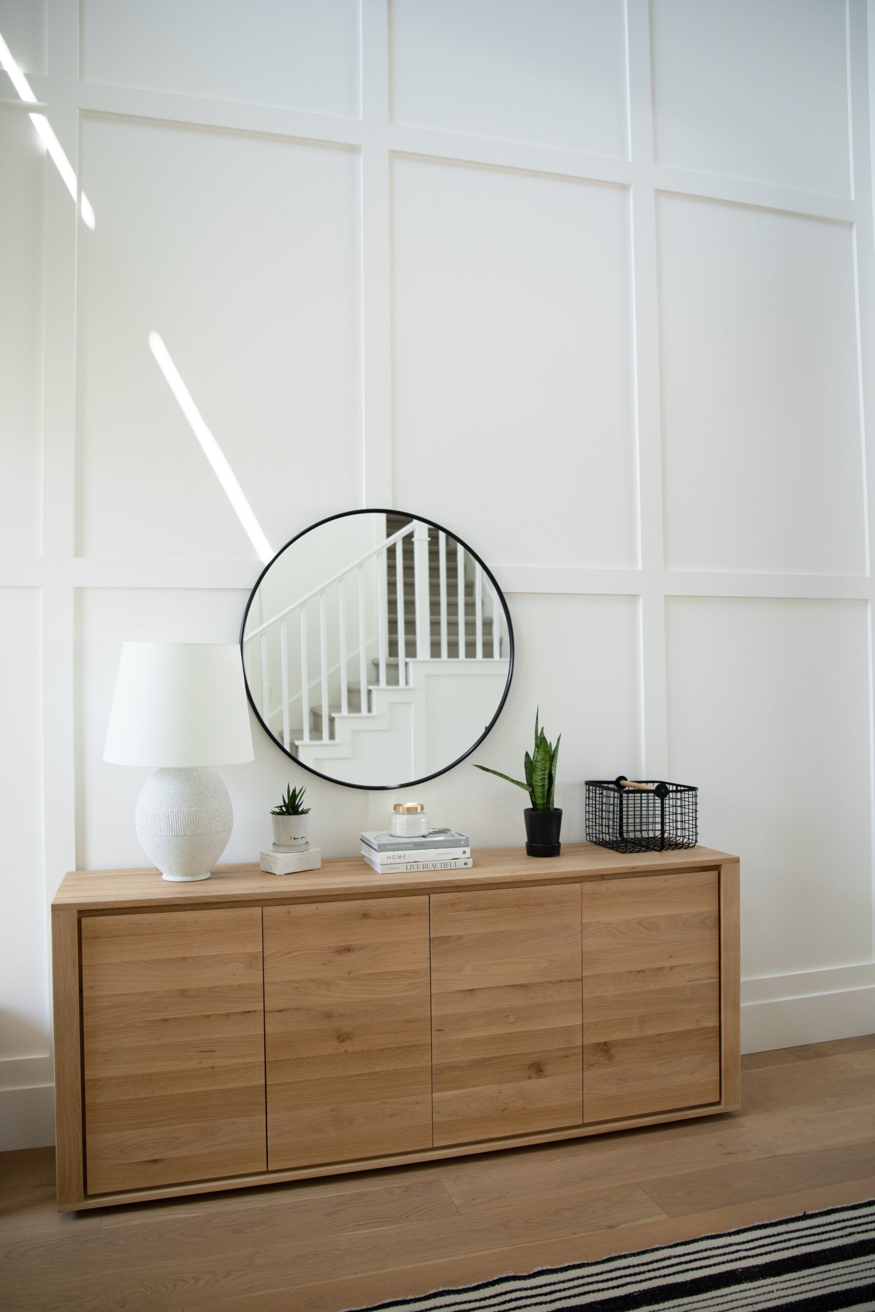 Light wood dresser with round mirror and lamp against white paneled wall in a bright room
