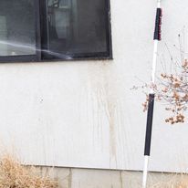 Snow pole beside a white house wall under a dark window, with dry grass at the base.
