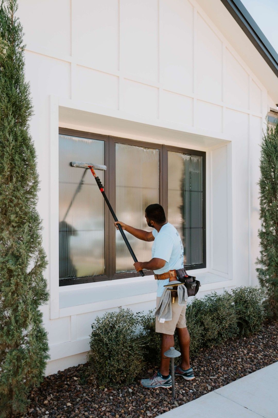 Person cleaning an exterior window with a squeegee beside hedges and a white building