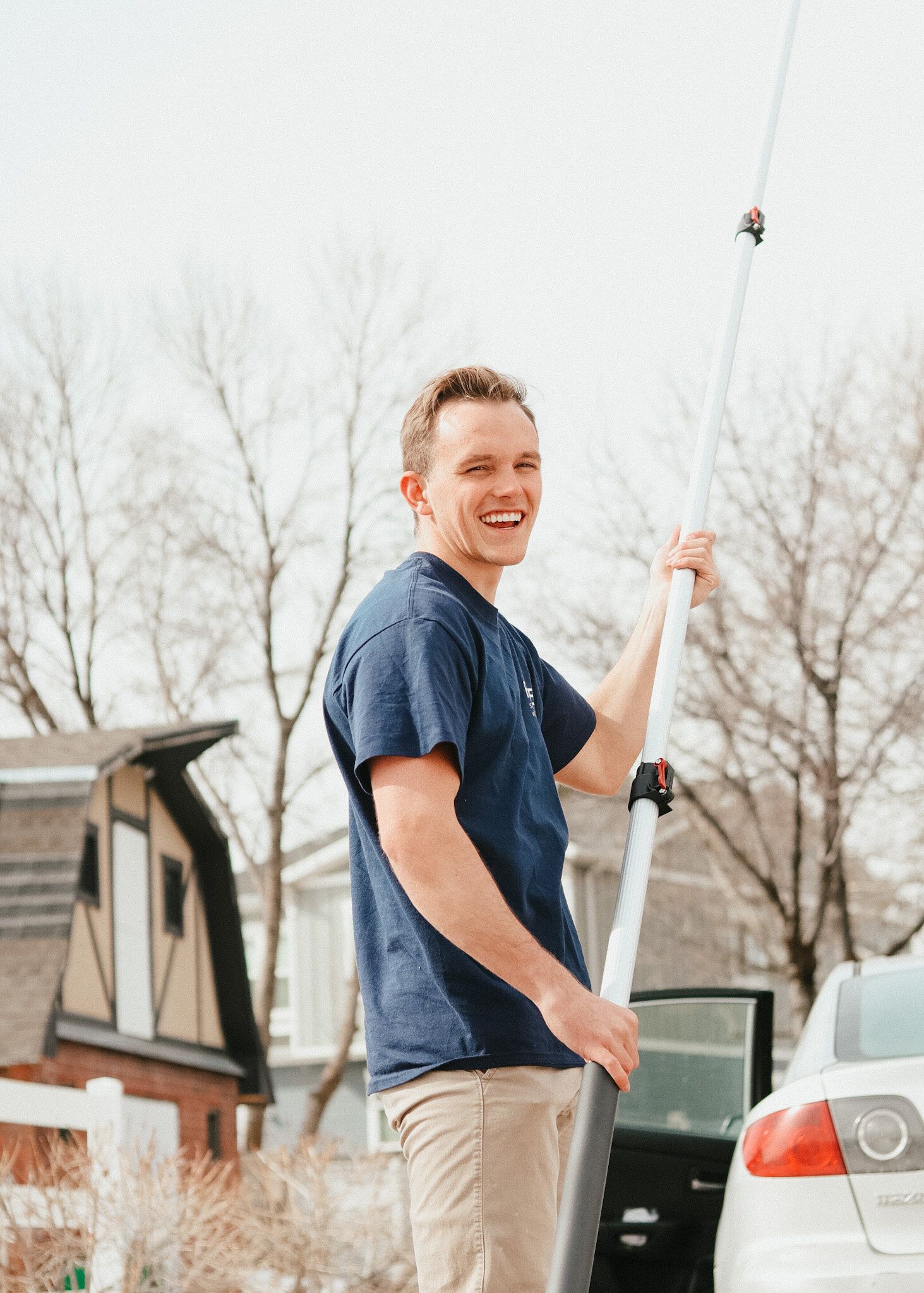 Person holding a long pole and smiling outdoors near parked cars and a house.