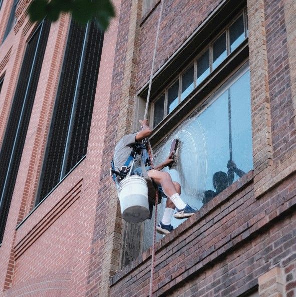 Window cleaner suspended on a brick building, cleaning an upper-story window with a bucket hanging beside him.