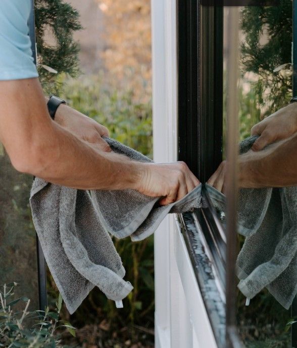 Person wiping a window with a gray cloth, with reflection in the glass.